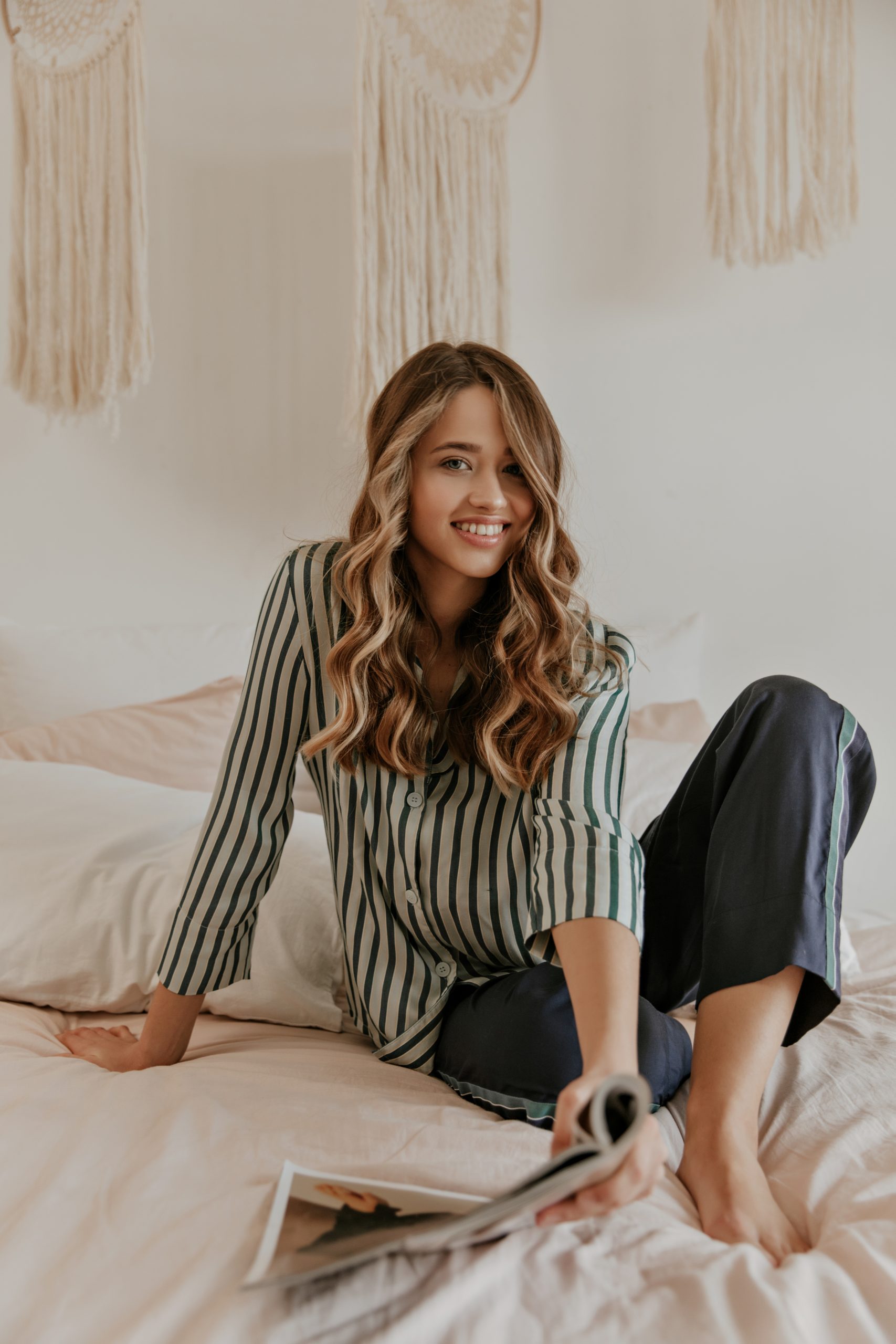 Charming lady in stylish striped shirt and silk pajama pants looks into camera, sits on soft beige bed and holds interesting magazine.