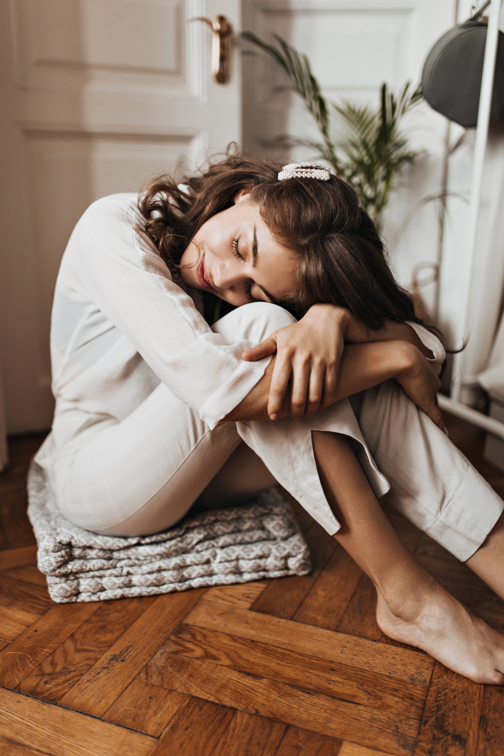 Woman sitting on wooden floor and sleeping. Pretty young girl in beige shirt and pants resting in cozy white apartment